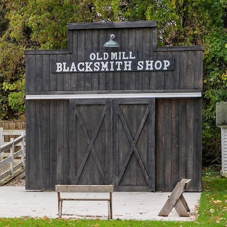 Black building with a sign that reads "Old Mill Blacksmith Shop". Two wooden benches sit in front of the building.