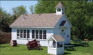 Old school house painted white with steeple. Separate bell steeple in front with an old hay rake.